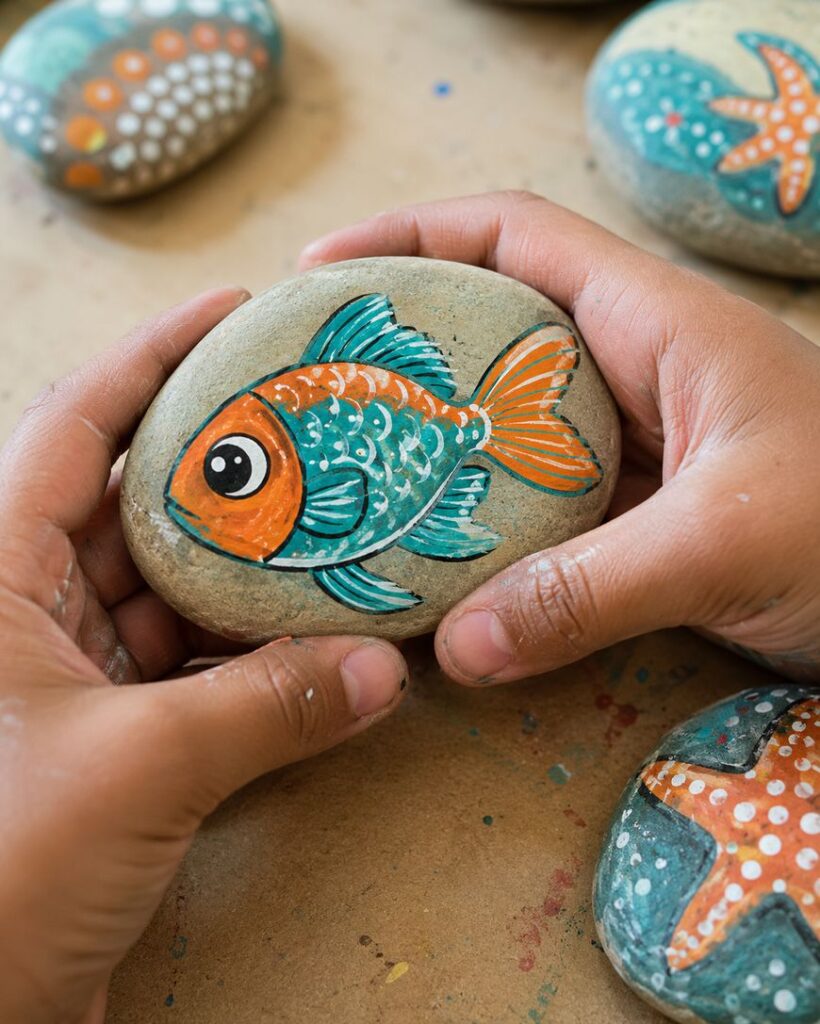 Two hands hold a painted rock featuring a colorful orange and blue fish, with other decorated rocks, including one with a starfish, visible on a brown surface in the background.