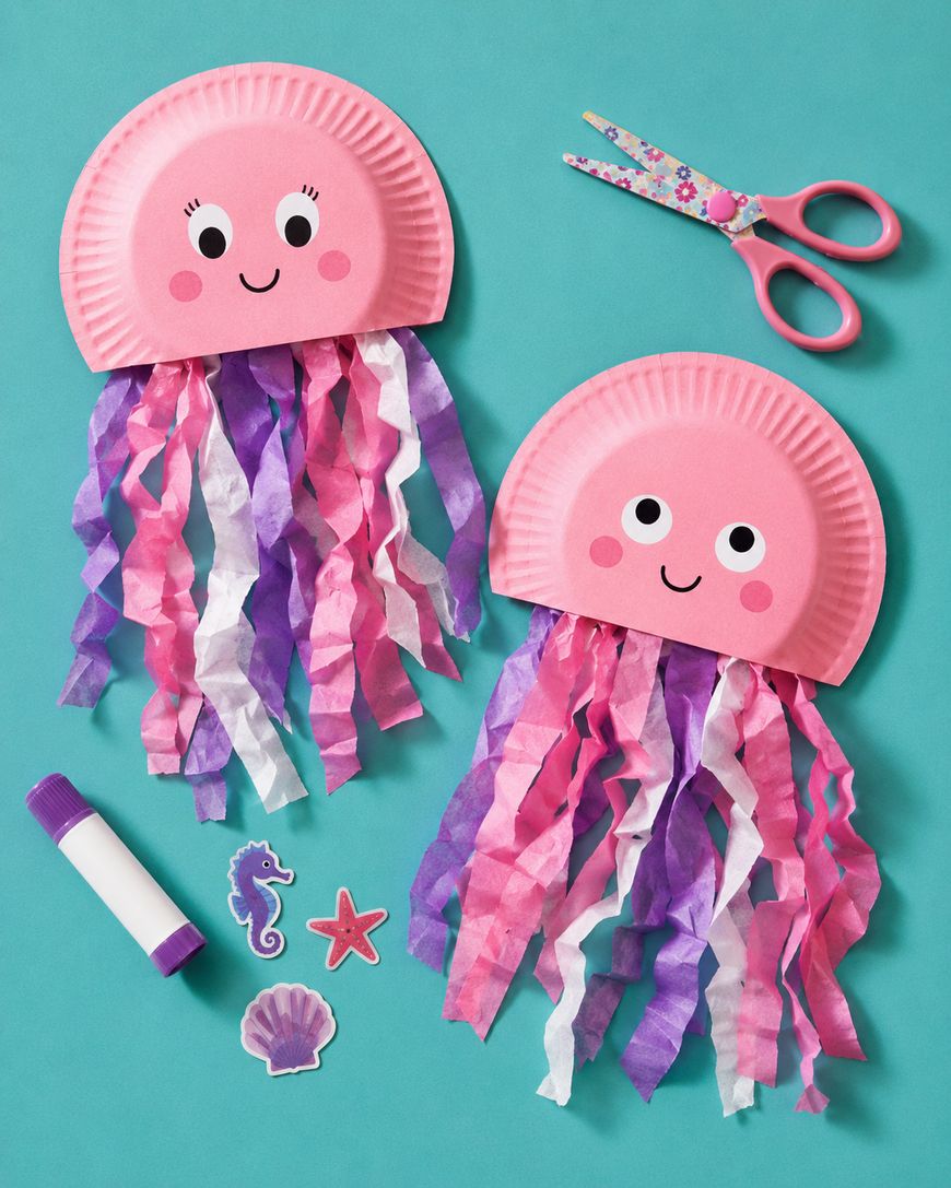 Two pink paper plate jellyfish crafts with smiling faces and tissue paper tentacles in pink, purple, and white. Nearby are scissors, a glue stick, stickers, and a purple seahorse sticker on a teal background.