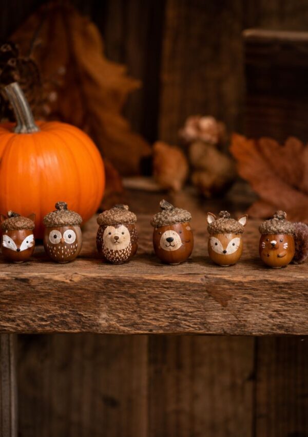Six acorns painted as woodland animals, including a fox, owl, hedgehog, bear, another fox, and a squirrel, sit on a rustic wooden surface with a pumpkin and autumn leaves in the background.