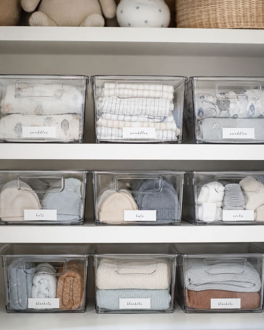 Clear plastic bins on white shelves, each labeled and organized with baby items: swaddles, hats, socks, and blankets in soft, neutral colors. Woven baskets and plush toys are visible on the top shelf.
