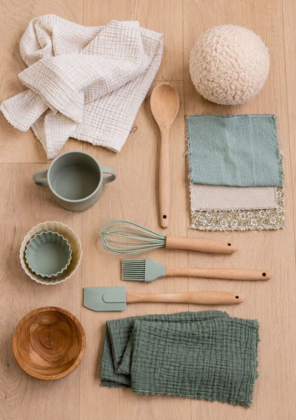 A flat lay of kitchen and home items on a wood surface, including cloths, a wooden spoon, silicone utensils, a mug, bowls, a whisk, a basting brush, and fabric swatches in neutral and green tones.