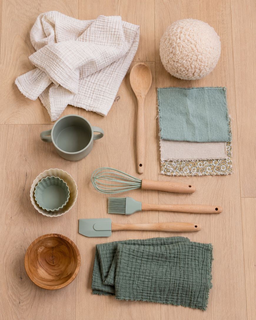 Various kitchen utensils and textiles, including cloths, a mug, a whisk, a basting brush, a spatula, a wooden spoon, a bowl, and fabric swatches, are arranged neatly on a light wooden surface.