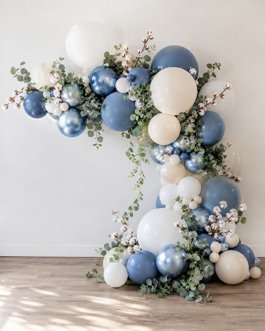A large balloon arrangement shaped like the number one, featuring blue, white, and cream balloons, decorated with green eucalyptus leaves and white flowers, against a white wall on a wooden floor.