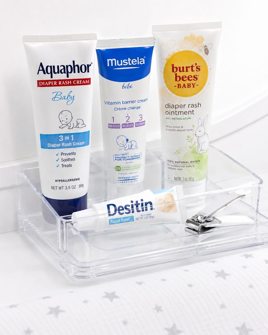 Four baby care products, including Aquaphor, Mustela, Burt’s Bees, and Desitin diaper creams, are arranged in a clear tray with a nail clipper, on a white surface decorated with grey stars.