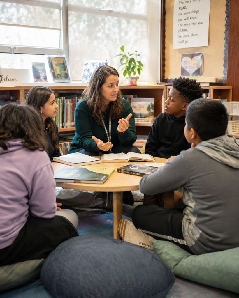 A teacher sits on the floor with four students around a small table in a classroom, engaging in discussion. Books are on the table, and bookshelves and educational decor are visible in the background.