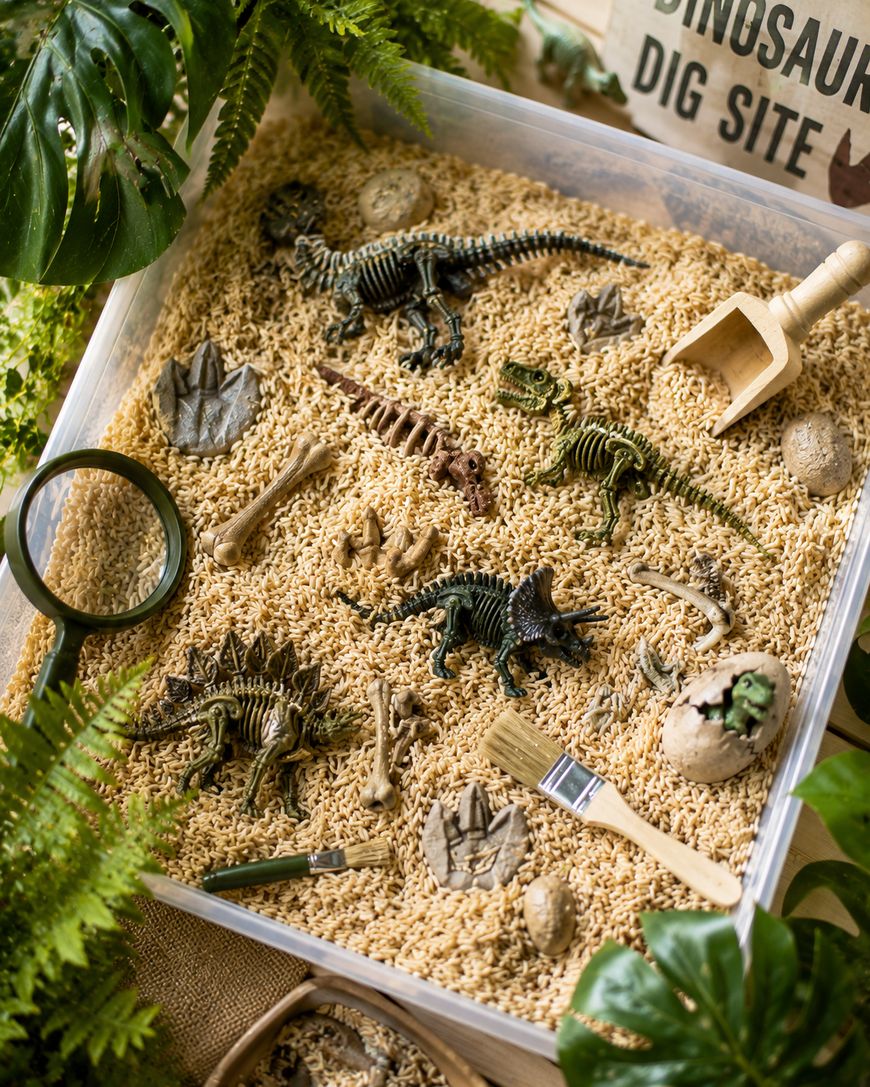 A dinosaur dig playset with toy skeletons, bones, eggs, sand tools, and brush in a sensory bin filled with rice, surrounded by green leaves and a magnifying glass.