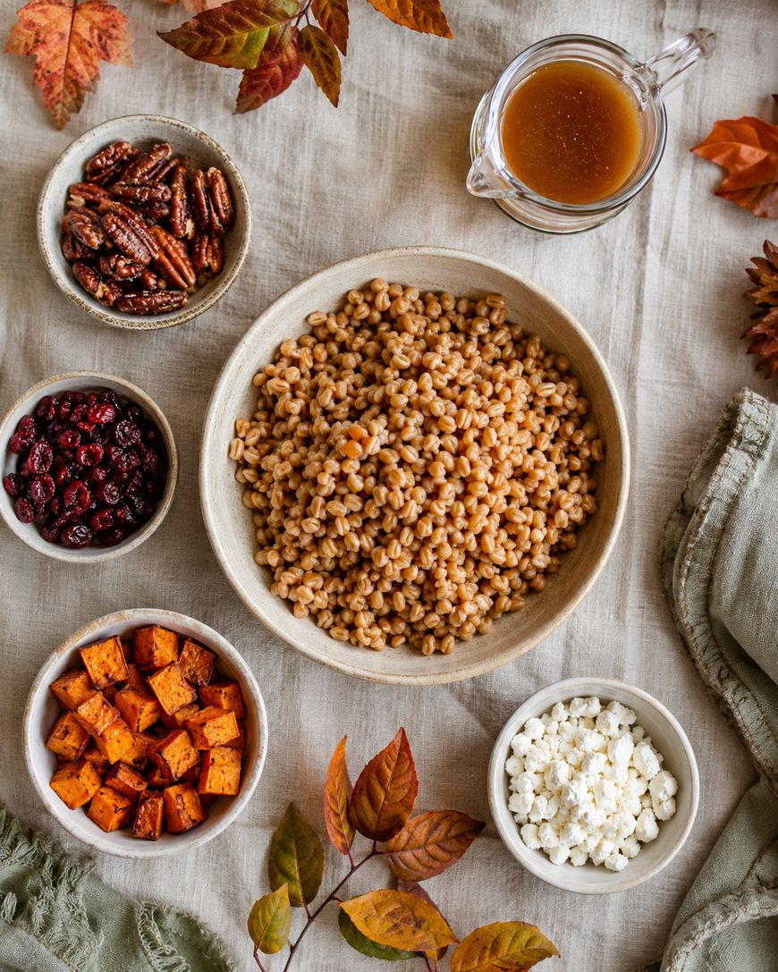 A fall-themed table setting with bowls of cooked grains, roasted sweet potatoes, dried cranberries, candied pecans, crumbled cheese, and a small pitcher of dressing, surrounded by autumn leaves on a beige cloth.