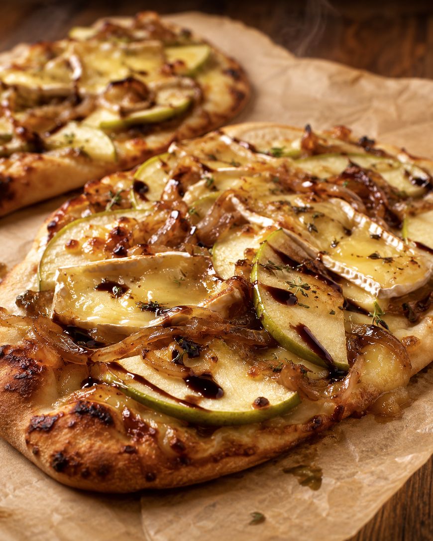 Flatbread pizza topped with melted cheese, thin green apple slices, caramelized onions, and a drizzle of balsamic glaze, served on parchment paper with another flatbread in the background.