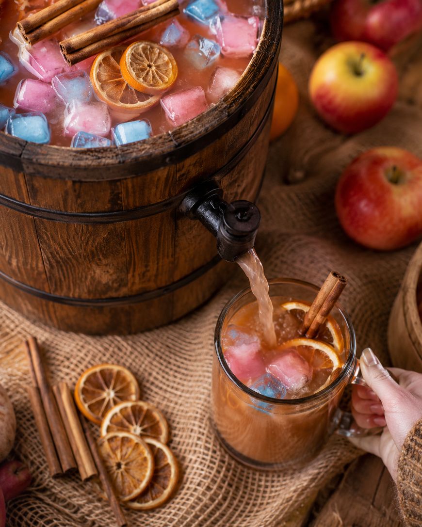 A wooden barrel dispenser pours a fruity drink with colorful ice cubes, dried orange slices, and cinnamon sticks into a glass mug. Apples, dried oranges, and cinnamon sticks are scattered on a burlap surface.