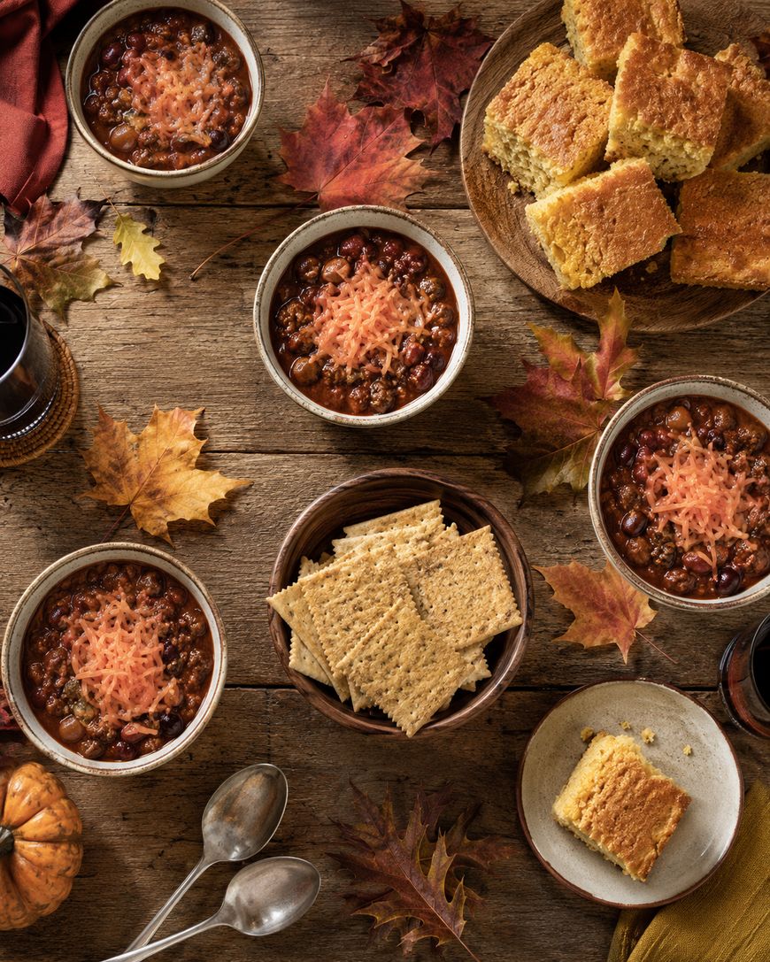 Four bowls of chili topped with cheese, plates of cornbread, and a bowl of crackers on a rustic table with autumn leaves, spoons, and drinks, creating a cozy fall meal setting.