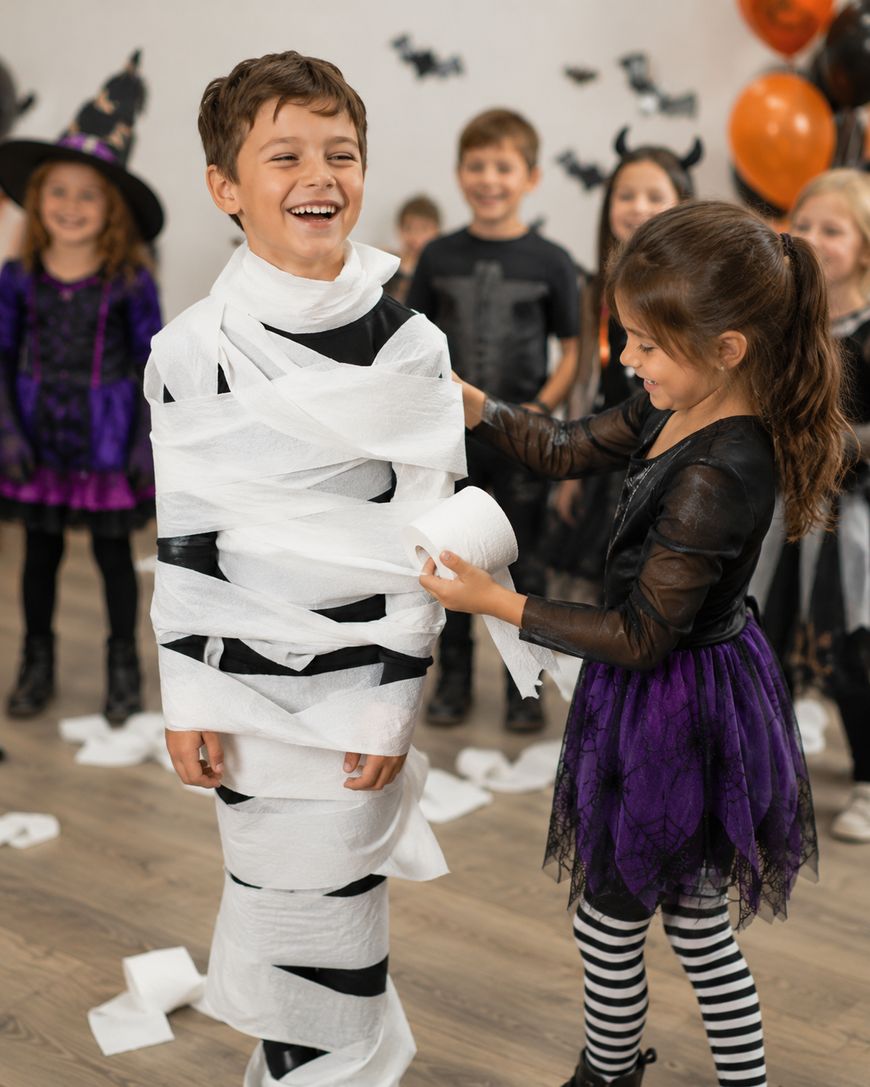 A smiling boy is being wrapped in toilet paper by a girl in a black and purple costume at a Halloween party. Other children in costumes and Halloween decorations are visible in the background.