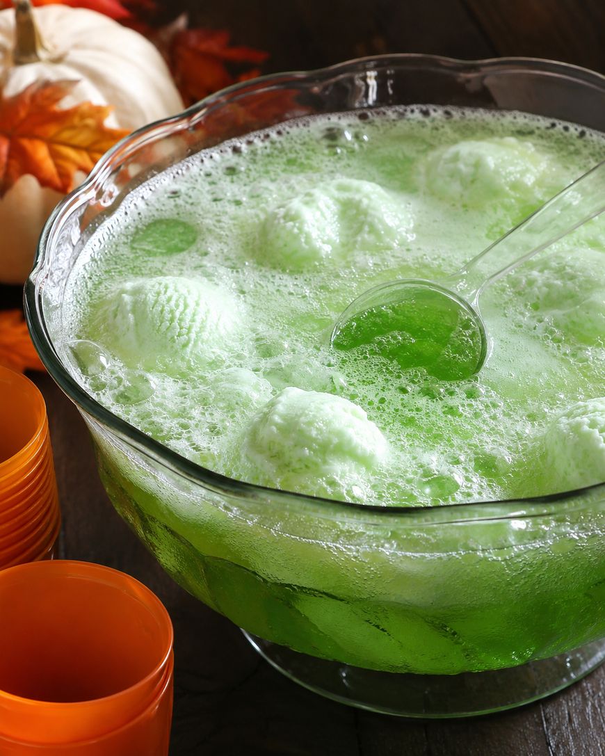 A glass bowl filled with foamy green punch and scoops of lime sherbet, with a ladle and orange cups nearby. Autumn leaves and a white pumpkin are in the background.