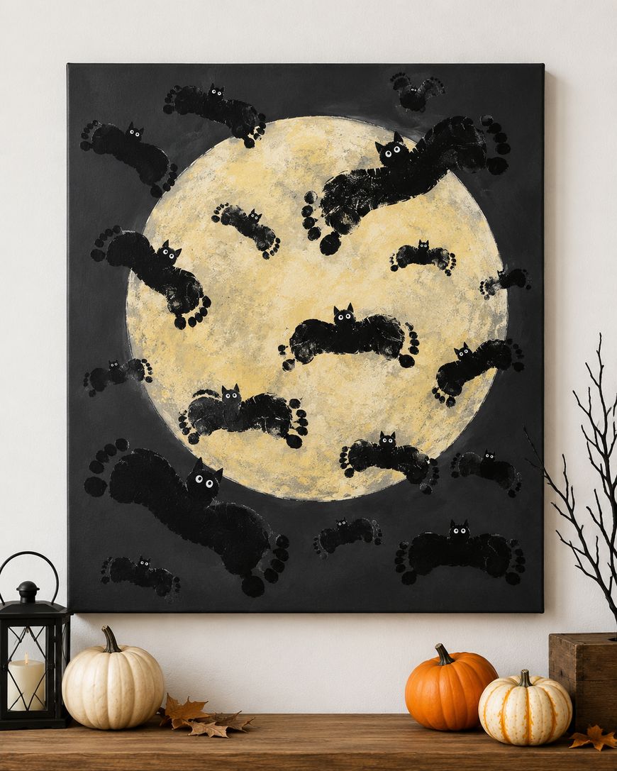 A painting of a large yellow moon on a black background, with black cat silhouettes made from painted footprints scattered across the moon and sky. There are pumpkins, a lantern, and leaves on the tabletop below.