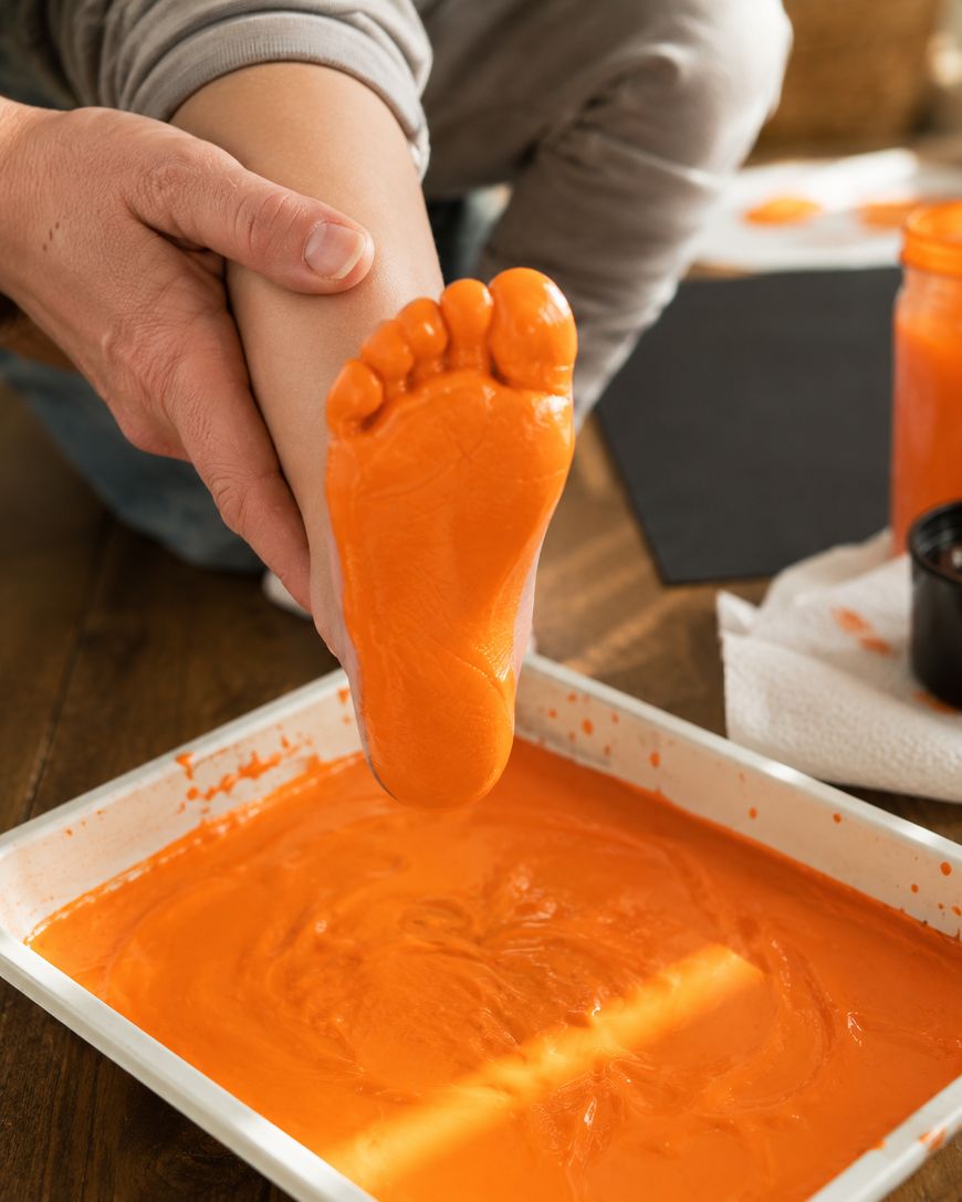 An adult holds a childs foot, which is covered in orange paint, above a tray filled with orange paint, likely preparing for a footprint art project.