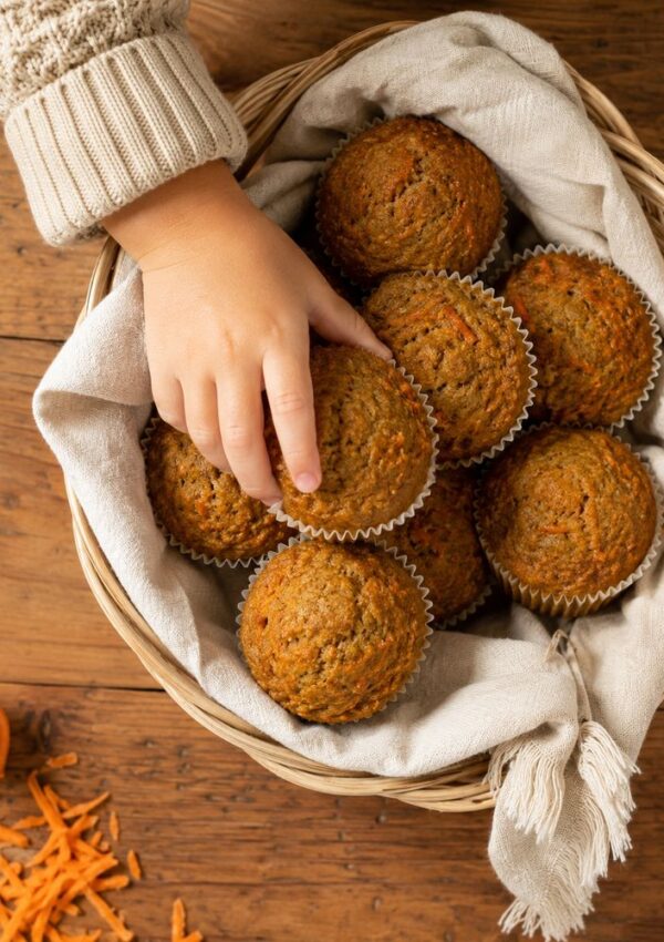 A child’s hand reaching for a muffin in a basket filled with muffins on a wooden table, lined with a beige cloth. Grated carrots are visible nearby.