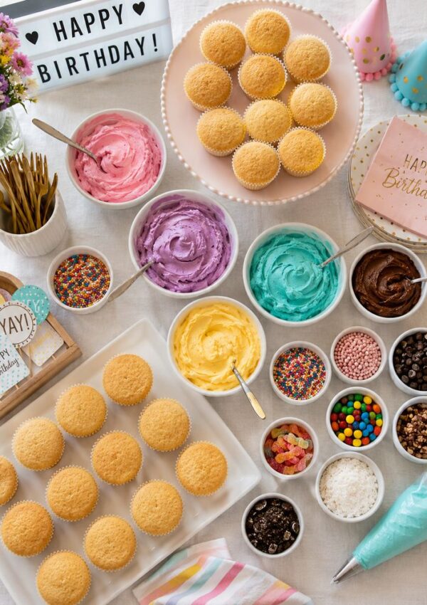 A birthday cupcake decorating setup perfect for fun baking activities, with plain cupcakes, colorful frosting, sprinkles, toppings, party hats, flowers, napkins, forks, and a “Happy Birthday” sign all neatly arranged on the table.