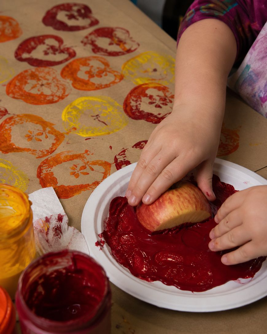 A child uses a halved apple dipped in red paint to stamp patterns onto brown paper. Various stamped prints in red, yellow, and orange are visible alongside jars of paint.