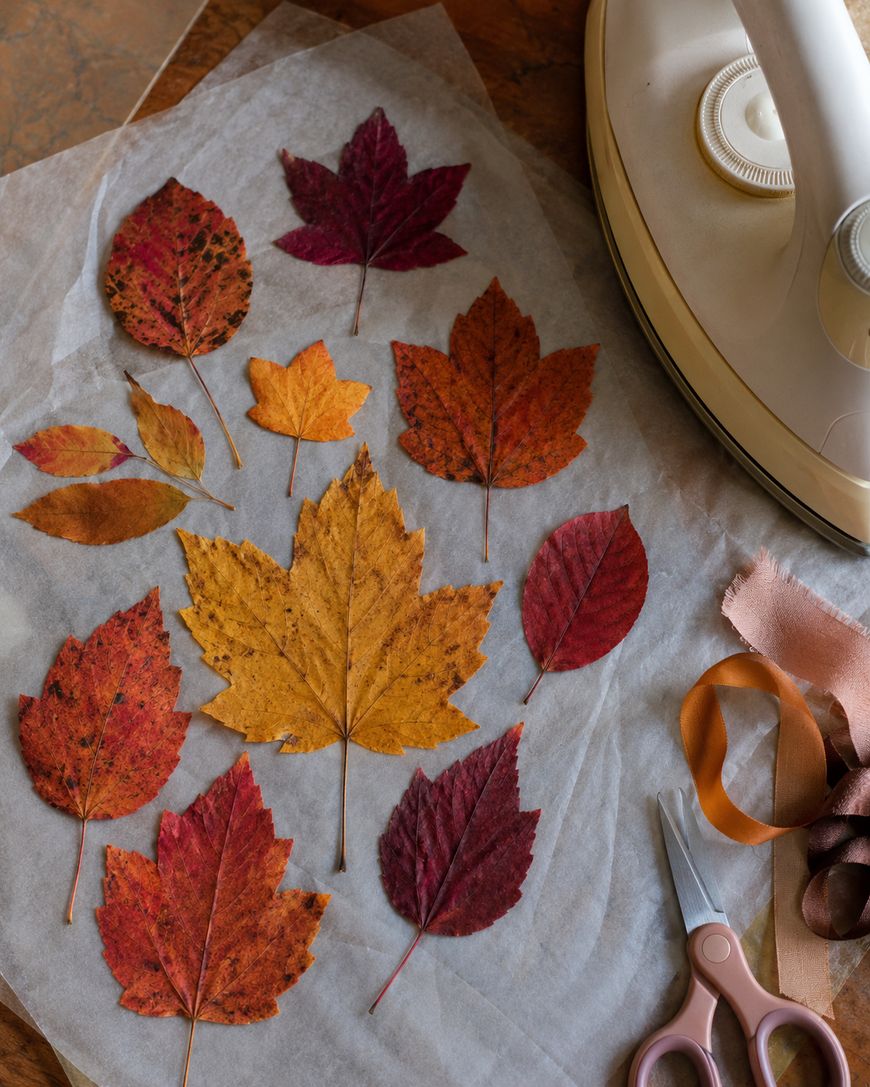 Colorful autumn leaves arranged on parchment paper beside an iron, scissors, and ribbon, suggesting a leaf-preservation or craft project on a wooden surface.