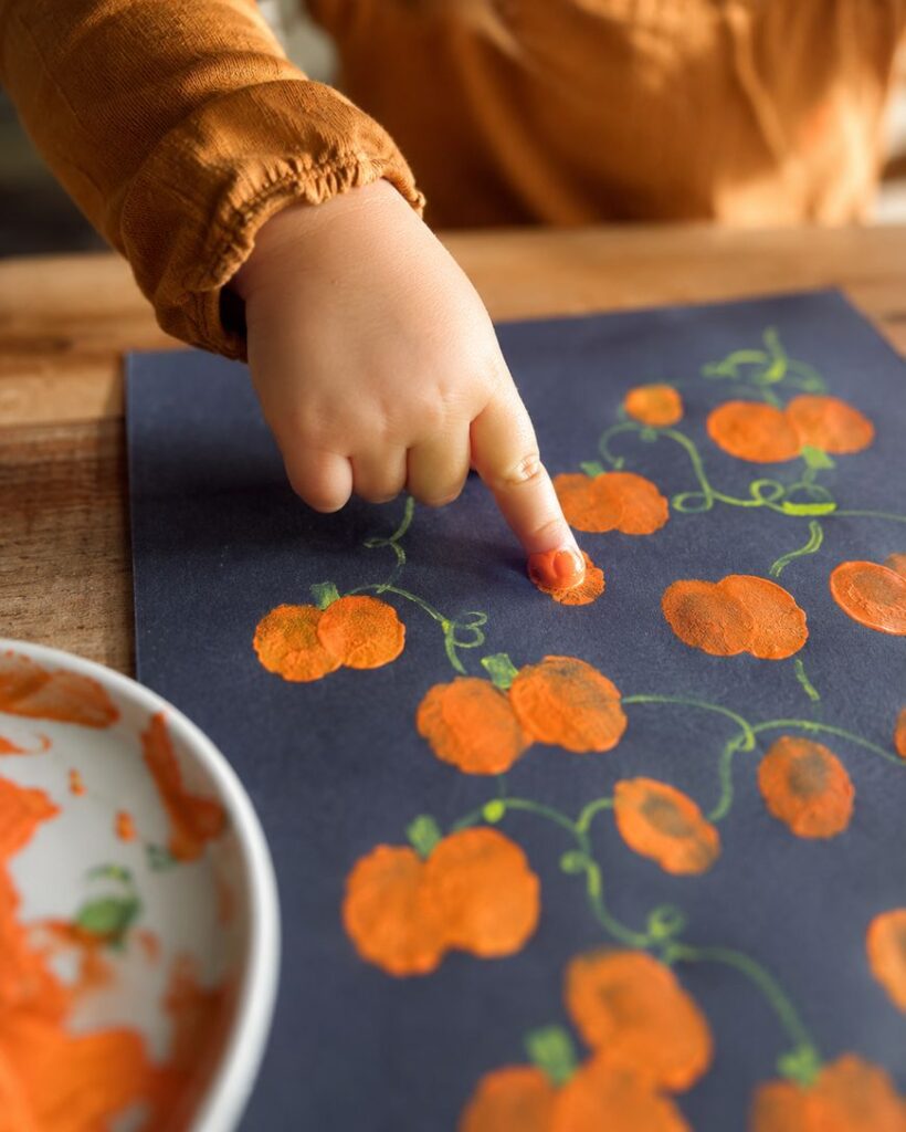 A child in an orange shirt uses their finger to paint pumpkin shapes with orange paint on black paper. A white plate with orange paint is nearby on the wooden table.