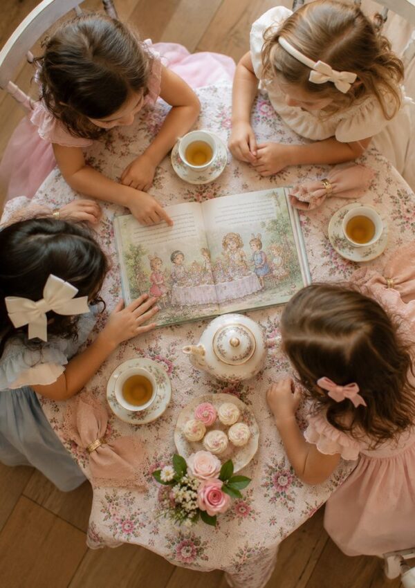 Four young girls in pastel dresses have a tea party, sitting around a table with tea cups, a teapot, cupcakes, flowers, and an open illustrated book. The table is covered with a floral tablecloth.