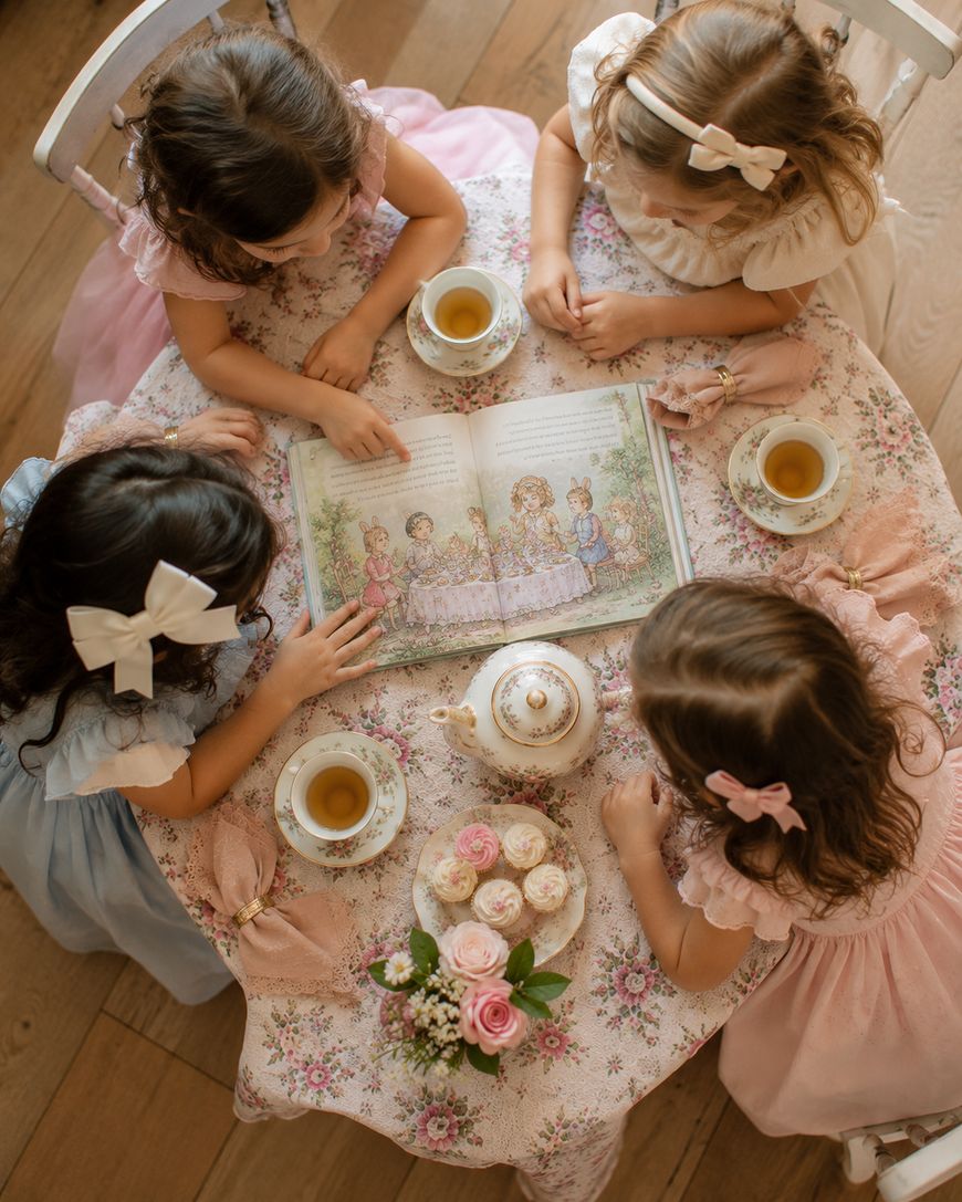 Four young girls in pastel dresses sit around a floral table, drinking tea and reading a book