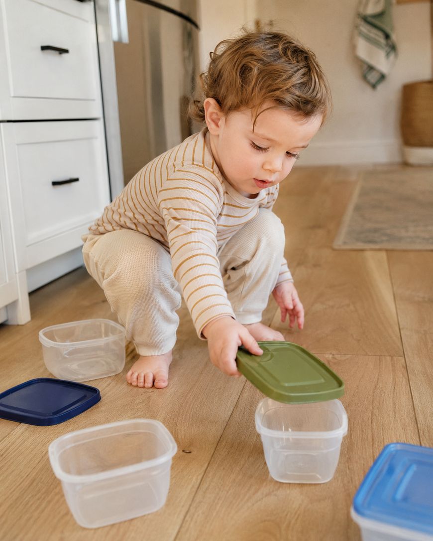 A toddler with curly hair crouches on a wooden floor, matching lids to plastic containers. Several lids and containers are scattered around, and the child focuses on placing a green lid on one container.