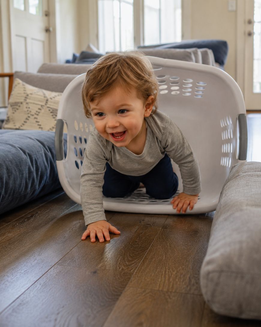 A smiling toddler crawls out of a white laundry basket on a wooden floor, surrounded by grey cushions in a cozy, sunlit living room.