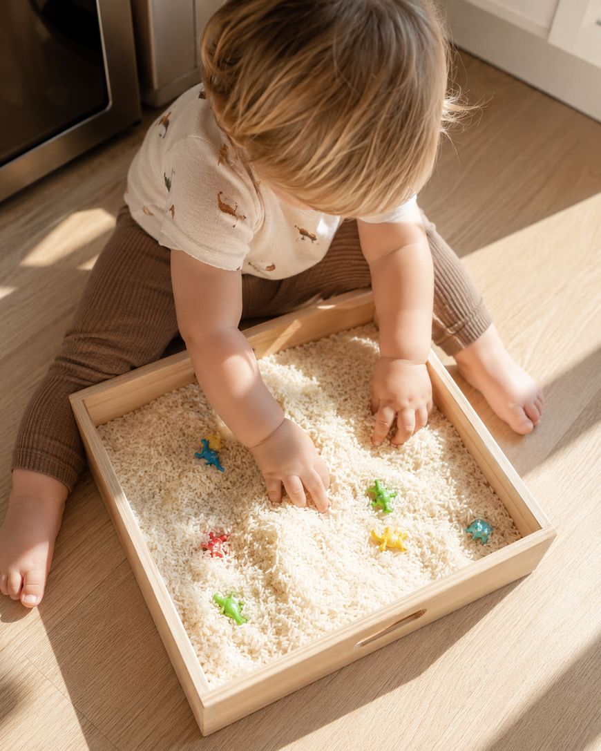 A young child sits on the floor, playing with a wooden tray filled with rice and colorful plastic animal toys, exploring textures with both hands.