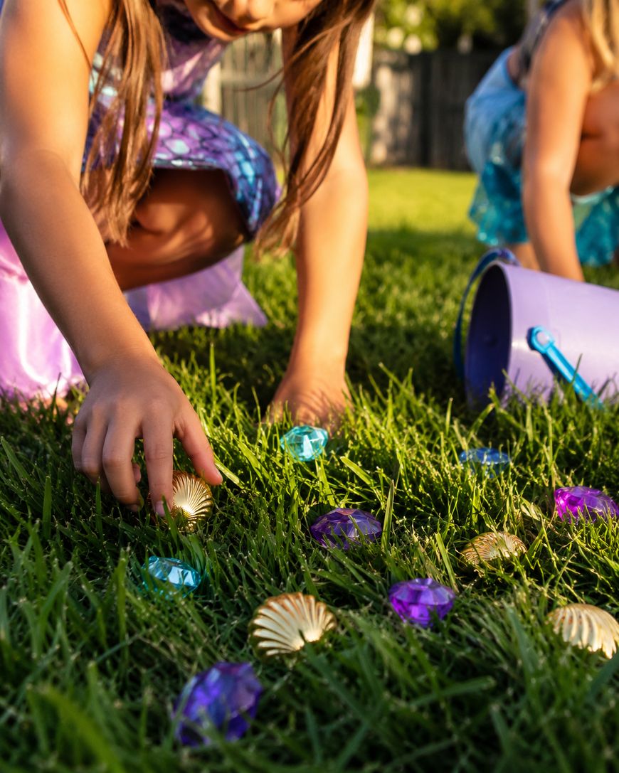 Two children in colorful dresses pick up toy gems and shell-shaped objects from green grass, with a tipped-over purple bucket beside them on a sunny day.
