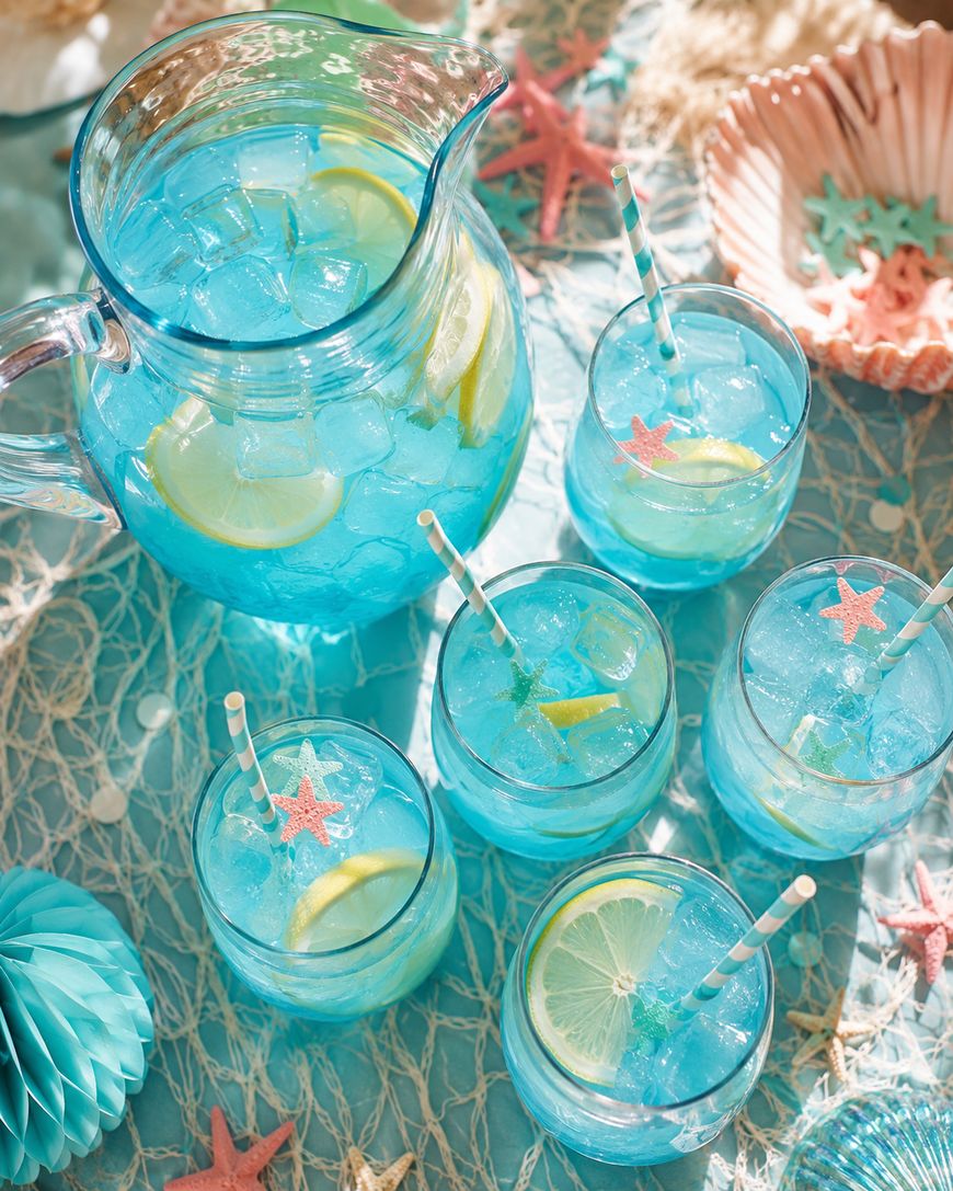 A pitcher and five glasses of blue lemonade with ice and lemon slices sit on a net-covered table, decorated with pastel starfish, seashells, and striped straws, evoking a beachy summer theme.