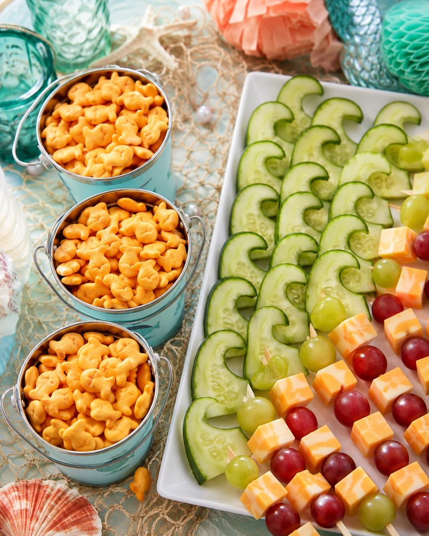 Three small buckets filled with goldfish crackers next to a tray with cucumber slices, green grapes, red grapes, and cheese cubes arranged in rows, creating a colorful snack display.