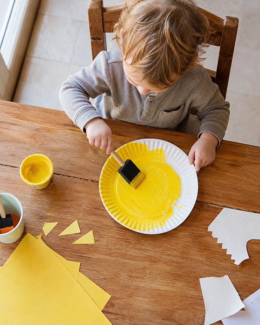 A young child sits at a wooden table, painting a white paper plate yellow with a foam brush. Art supplies, including yellow paper and a cup, are scattered around.
