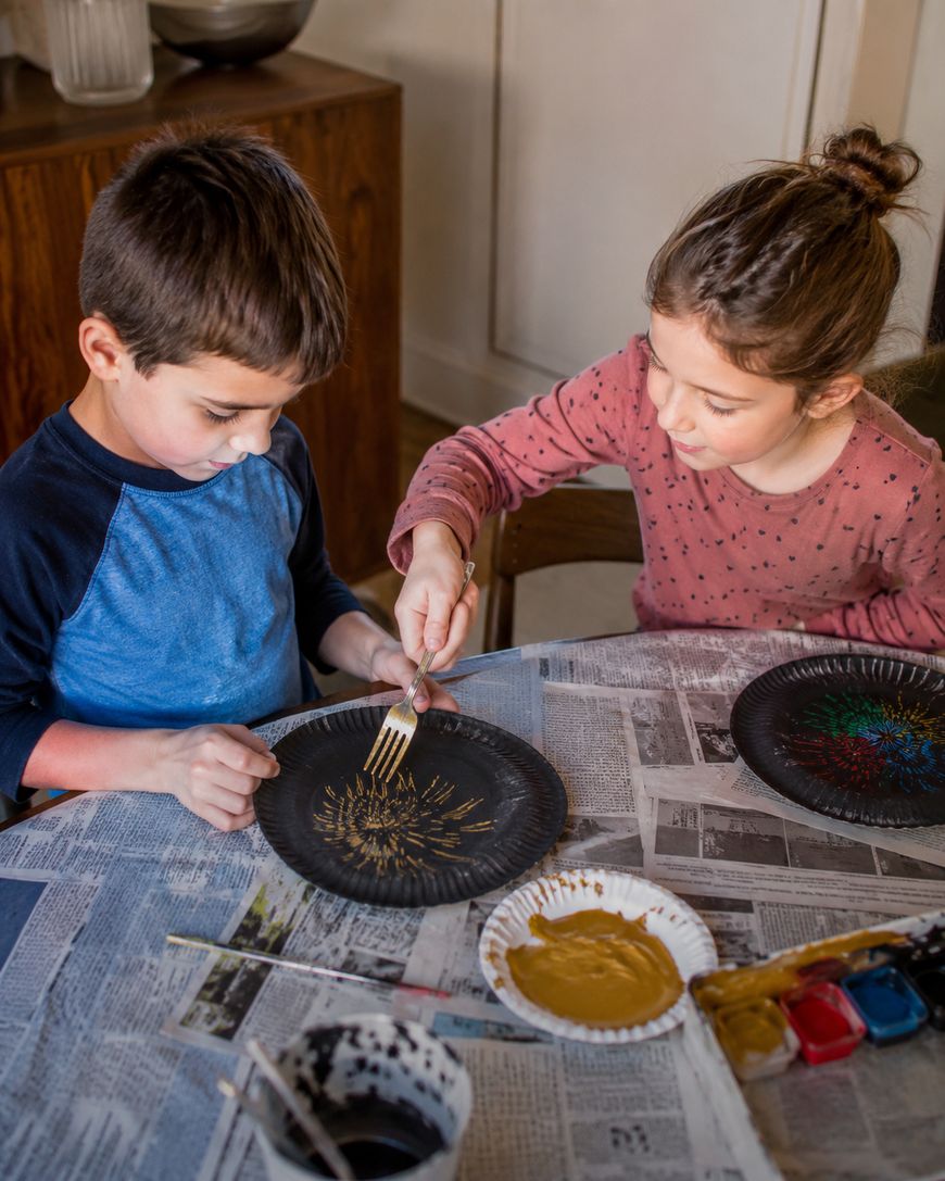 Two children sit at a table covered with newspaper, using forks to paint patterns on black paper plates. Paint, forks, and art supplies are visible on the table.