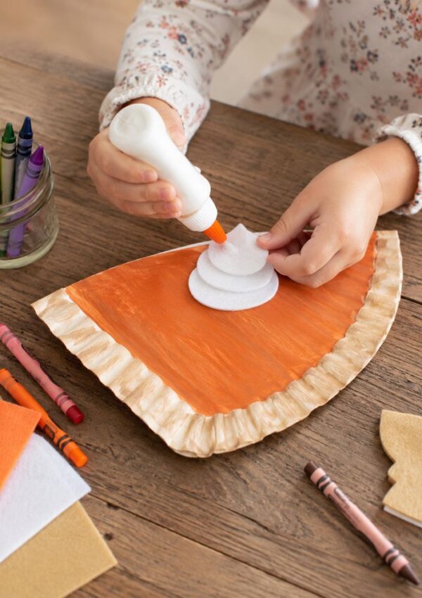 A child glues white felt onto an orange, pie-shaped craft made from a paper plate. Crayons, felt sheets, and a jar of crayons are nearby on a wooden table.