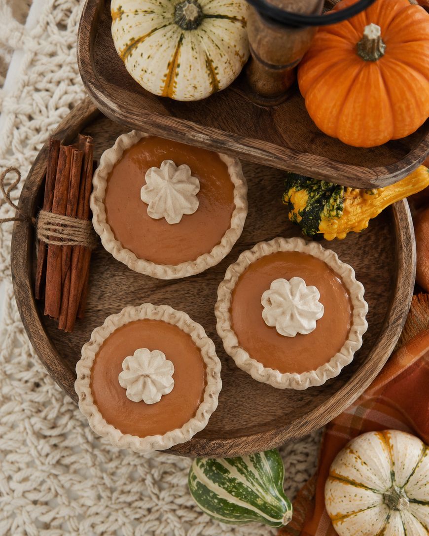 Three mini pumpkin pies with whipped cream on top are arranged on a wooden tray, surrounded by small decorative gourds and pumpkins, with a bundle of cinnamon sticks nearby.