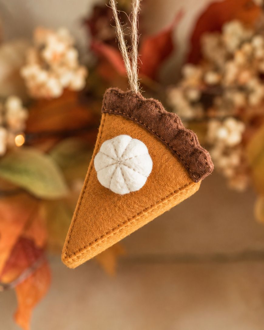 A felt ornament shaped like a pumpkin pie slice with a white whipped cream dollop, hanging by a string. Autumn leaves and flowers are blurred in the background.