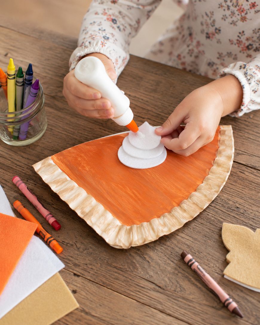 A child glues a white felt swirl onto a paper plate craft shaped and painted like a slice of pumpkin pie. Crayons, felt sheets, and a jar of crayons are on the wooden table.