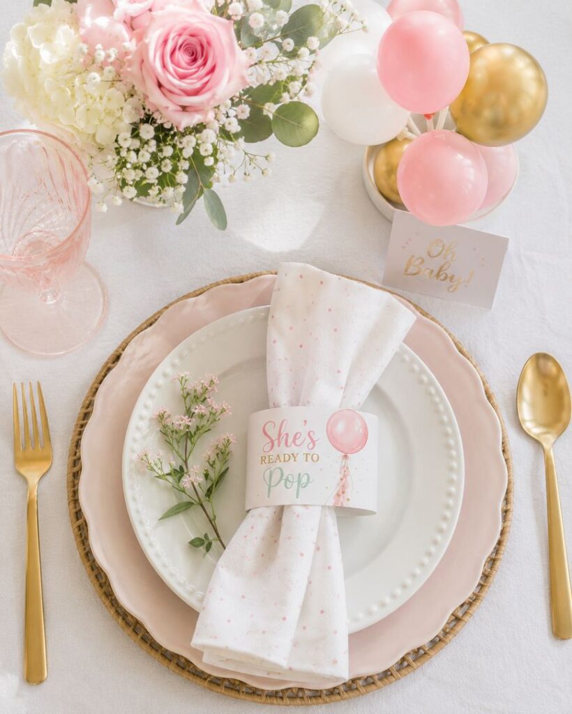 A baby shower table setting with pink and gold accents, a floral arrangement, gold cutlery, a “She’s Ready to Pop” napkin holder, balloons, and an “Oh Baby!” card on a white tablecloth.