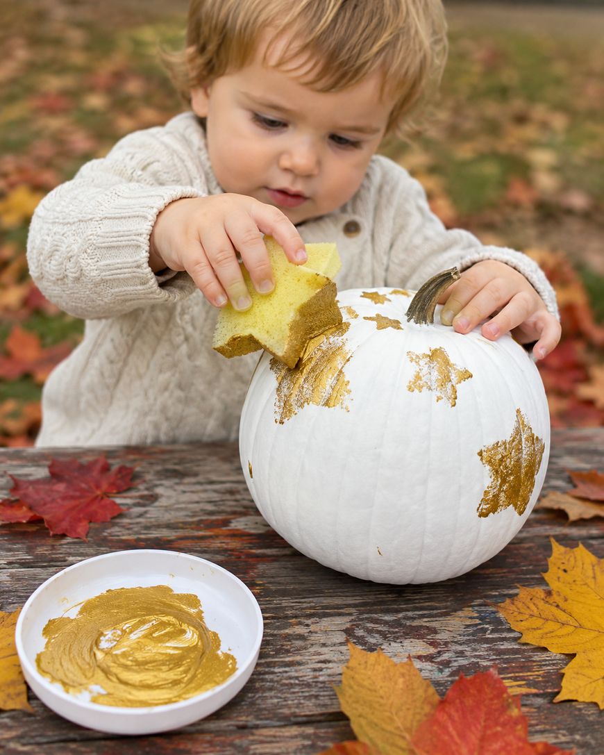 A young child uses a sponge to paint gold stars on a white pumpkin. A bowl of gold paint sits on a wooden table covered in autumn leaves.