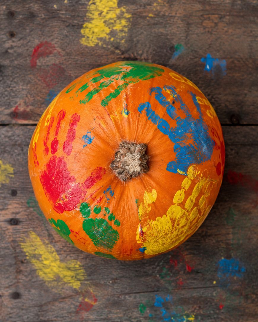 A pumpkin sits on a wooden surface, decorated with colorful handprints in red, yellow, green, and blue paint. Smudges of similar paint are visible on the wood around the pumpkin.