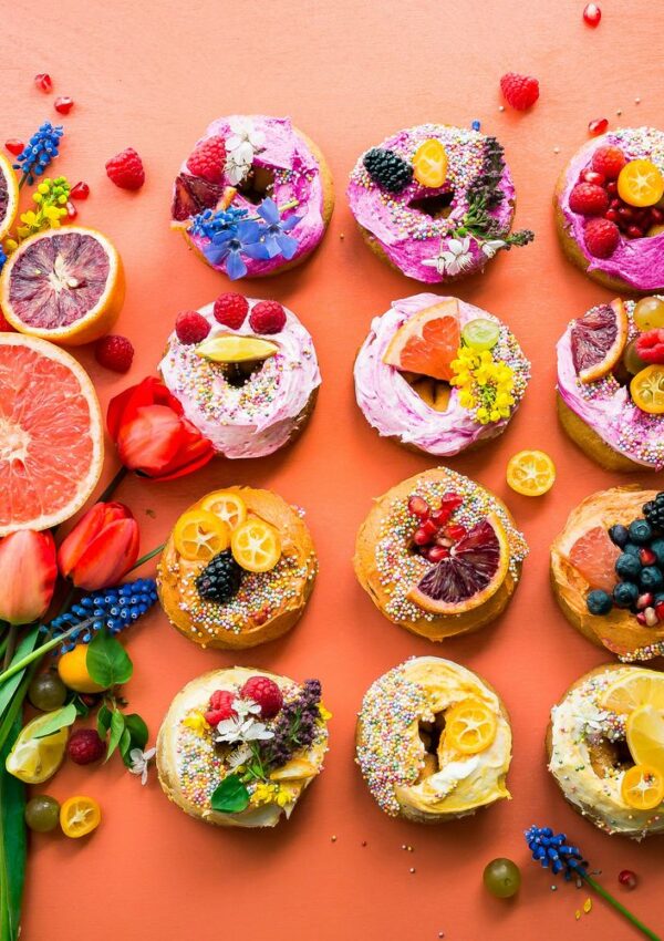 Twelve colorful donuts with vibrant frosting, fruit slices, berries, sprinkles, and edible flowers on a peach background, surrounded by citrus fruits and flowers.