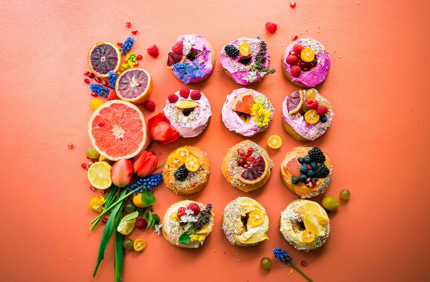 Colorful donuts topped with fruit, flowers, and sprinkles are arranged in a grid on an orange surface, surrounded by sliced citrus fruits, grapes, and flowers.