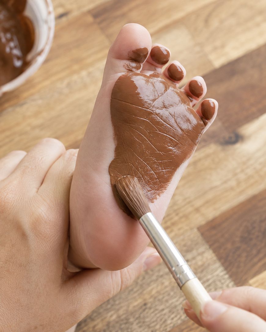 A hand uses a brush to paint the sole of a bare foot with brown paint. The scene is set on a wooden surface.