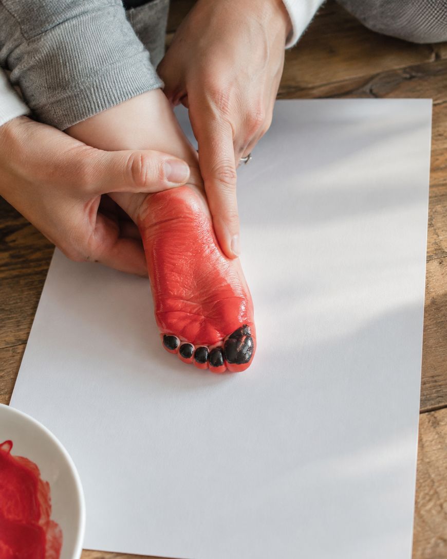 An adult presses a childs foot, painted red and black, onto a blank white paper to make a footprint, with a plate of paint nearby on a wooden surface.
