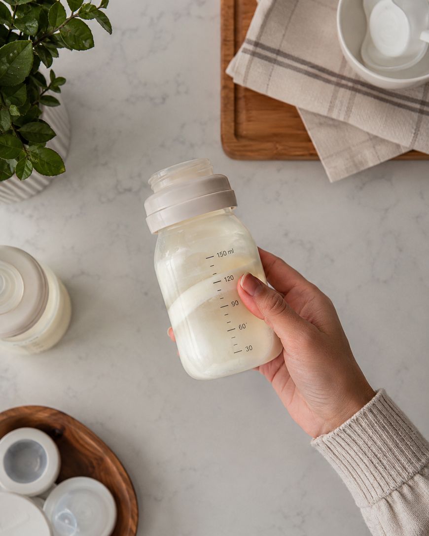 A hand holds a baby bottle filled with formula, next to a plant, wooden tray with cloth and bowls, and formula container on a white countertop.