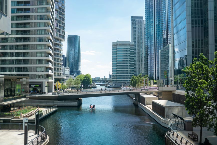 A modern cityscape with tall glass buildings, a calm canal, a pedestrian bridge, and a small boat on the water under a blue sky with a few clouds.