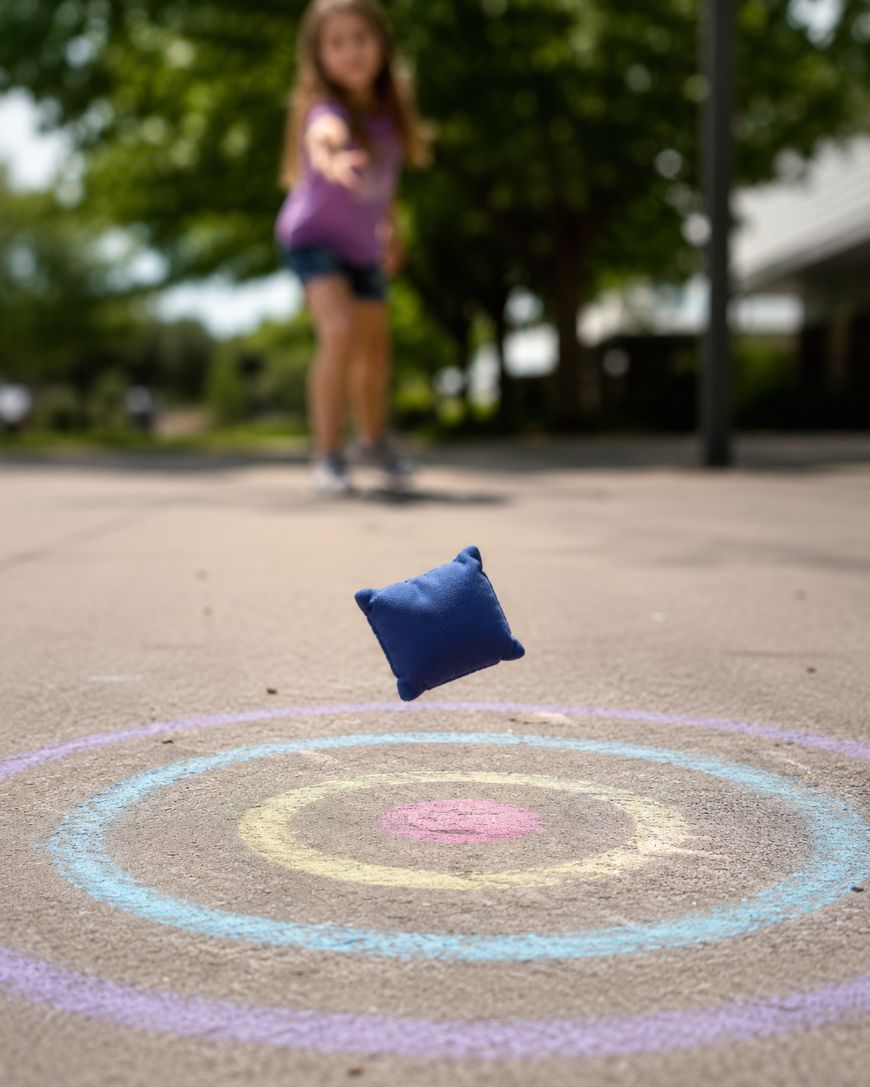 A girl throws a blue beanbag towards a chalk target drawn on the pavement.