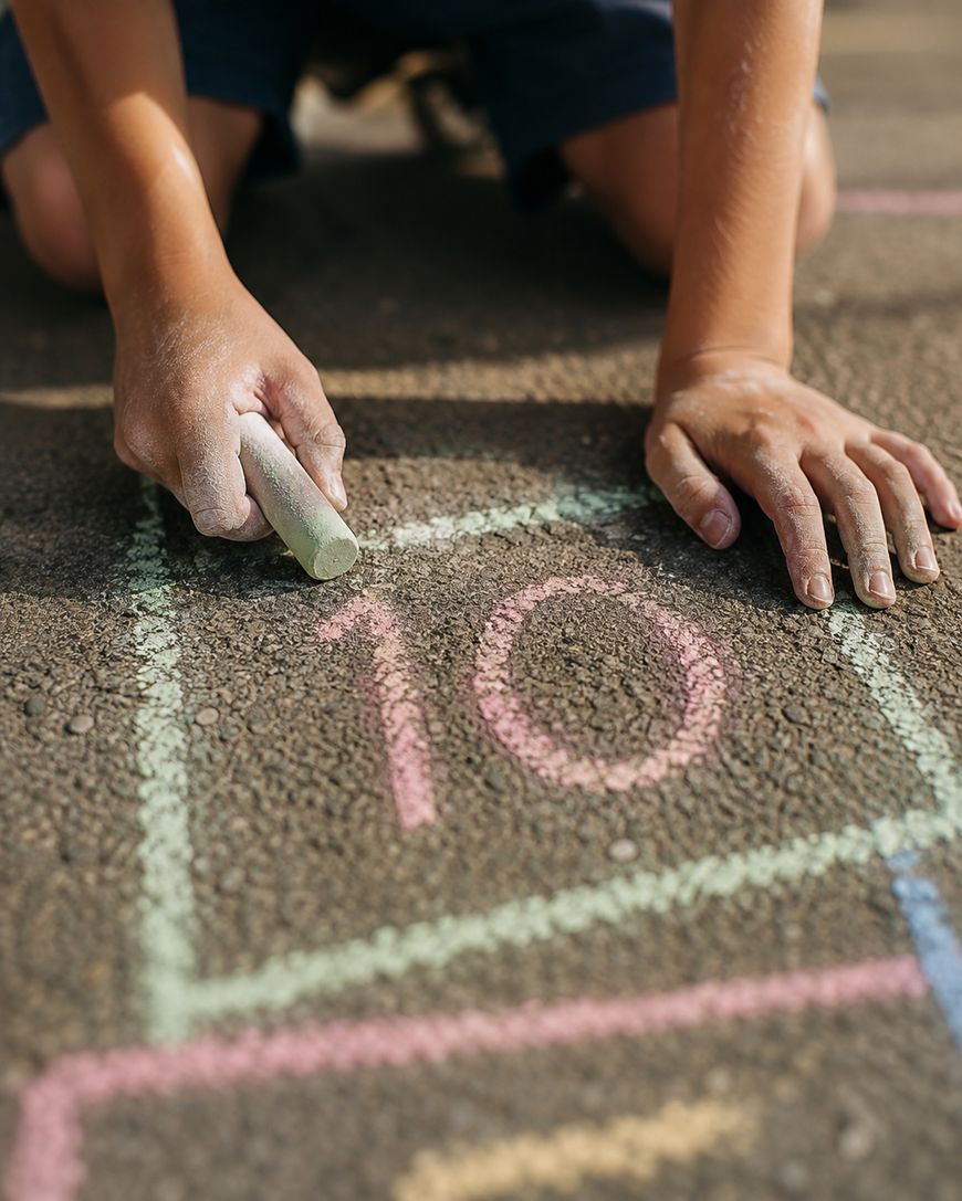 A child draws a hopscotch grid with colored chalk on asphalt, reaching the number 10.