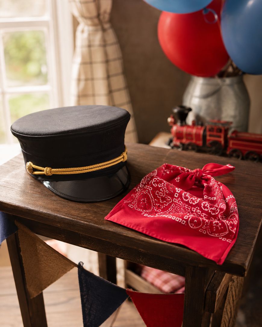 A black conductors hat and a red bandana sit on a wooden table, with a toy train and red and blue balloons in the background, near a window with checked curtains.