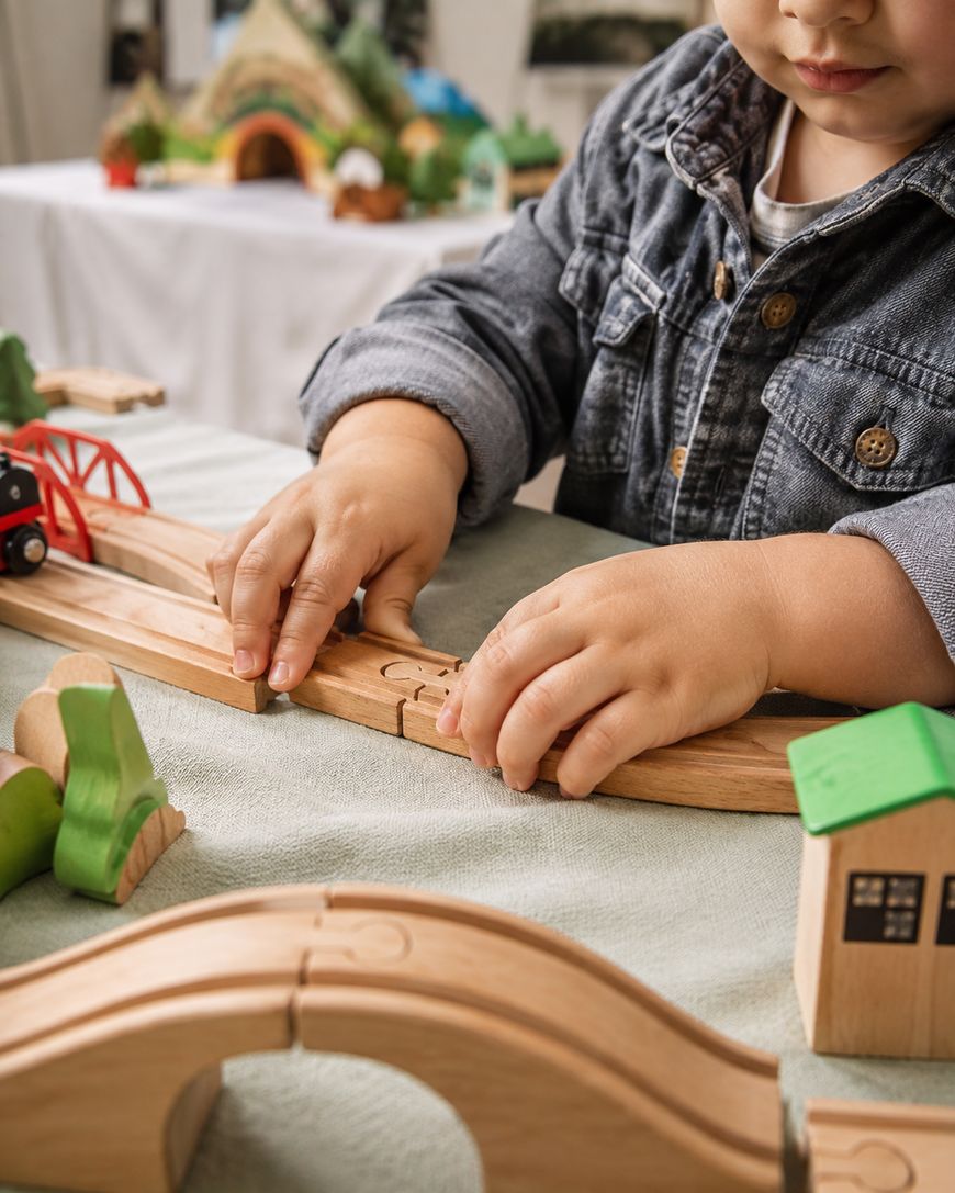 A young child in a denim jacket plays with a wooden train set on a table, arranging tracks and bridges with their hands.